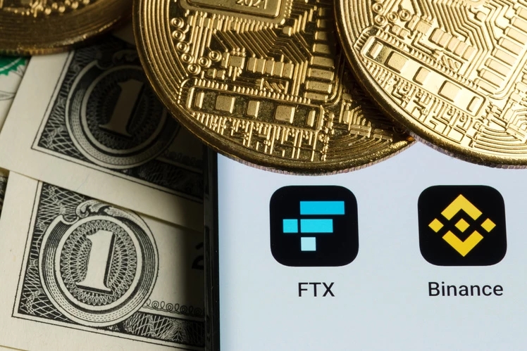 une photo de billets de dollars américains, de pièces d'or en bitcoin et des applications iPhone de FTX et Binance représentant l'échec de l'opération d'acquisition de FTX