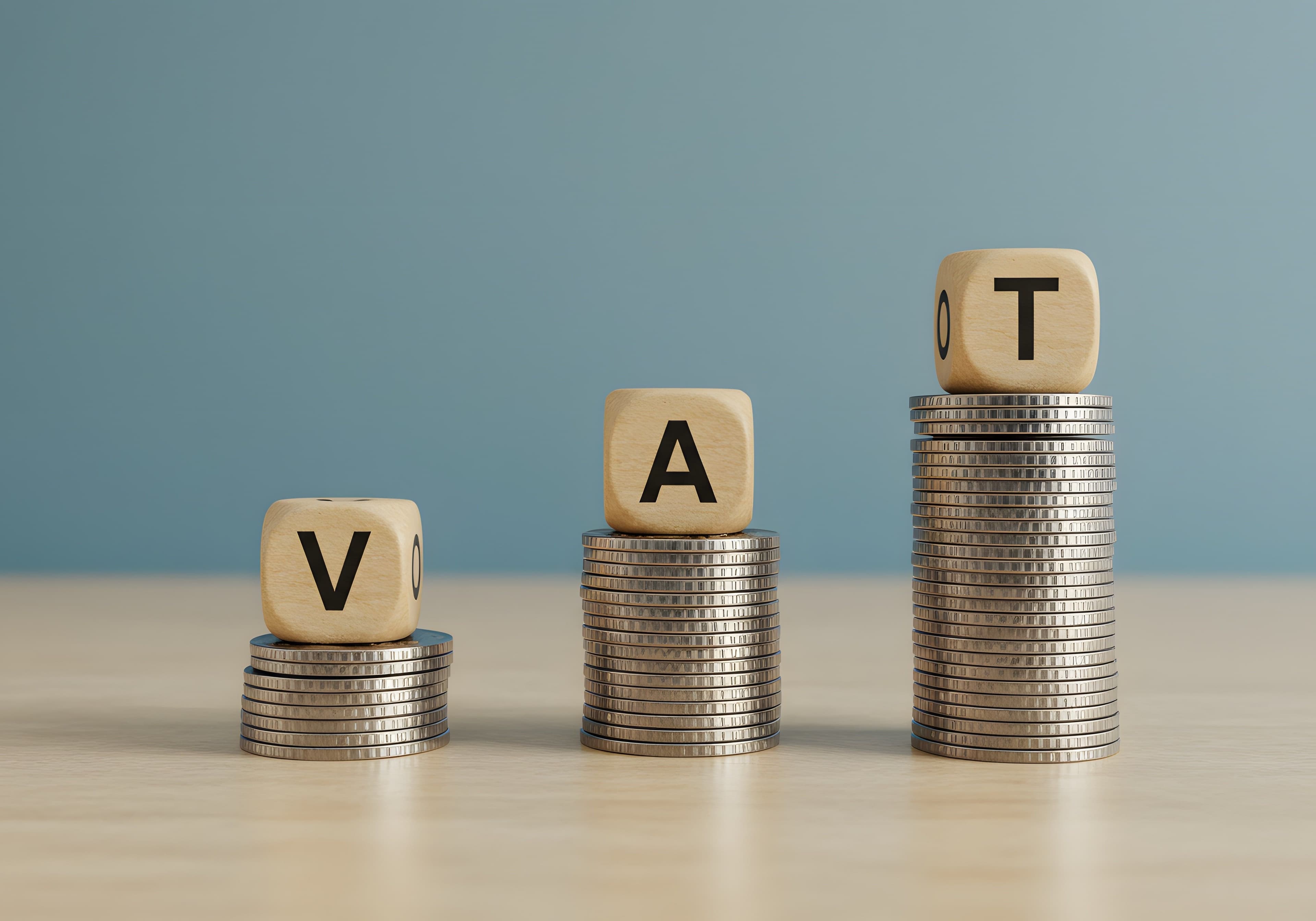 Holzwürfel mit den Buchstaben „V“, „A“ und „T“ auf gestapelten Münzen, die ansteigend angeordnet sind, vor blauem Hintergrund. Holzwürfel mit den Buchstaben „V“, „A“ und „T“ auf gestapelten Münzen, die ansteigend angeordnet sind, vor blauem Hintergrund.